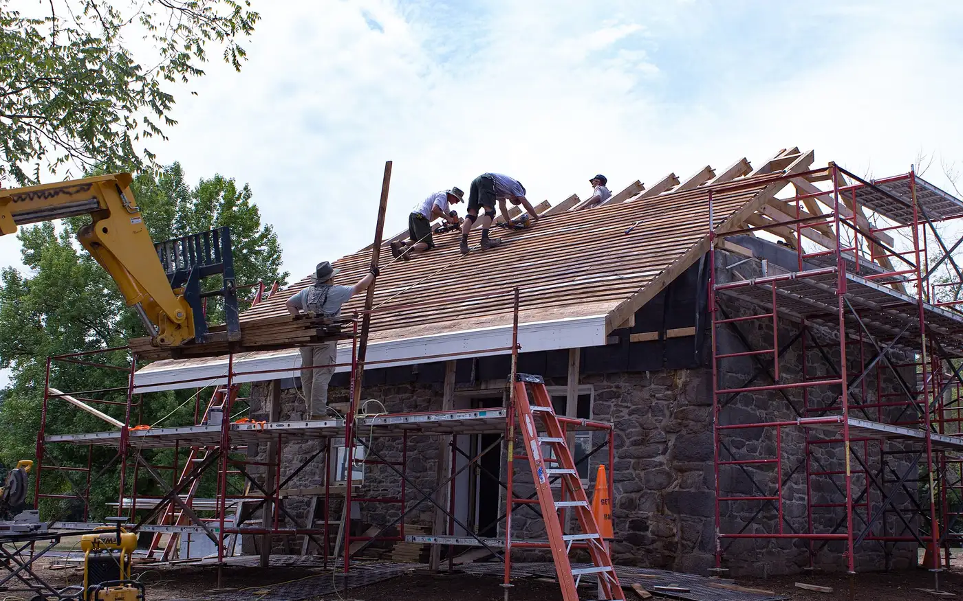 A roofing crew near the ridge as cedar shingles are lifted onto the roof by telehandler.